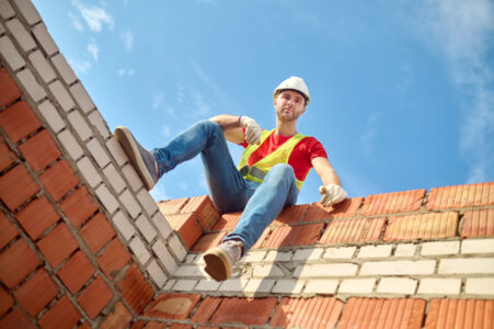 Short break. Bottom view of man in protective helmet and gloves sitting on brick wall looking at camera on sunny day at construction site